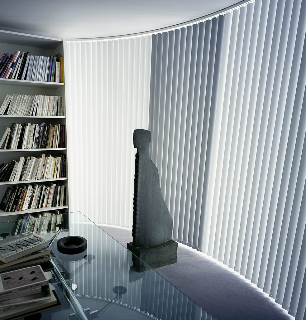 Ein Raum mit einem runden Glastisch, einem Bücherregal voller Bücher und einer abstrakten Skulptur vor vertikalen Jalousien.